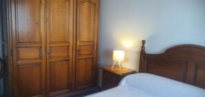Un dormitorio con un armario de madera, una mesita de noche con lámpara y una cama con edredón blanco.