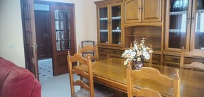 Un comedor con una mesa de madera, sillas y un jarrón de flores. Se observa un aparador de madera con puertas de cristal y un sofá rojo al fondo.