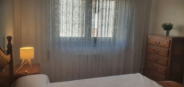 A room with a bed, a wooden dresser, a lamp, and a window with patterned curtains.