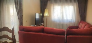 Una habitación con una televisión, controles remotos y una lámpara sobre una mesa de madera. Hay dos sofás rojos y una silla de madera.