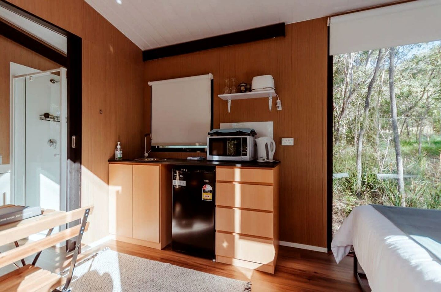 Une cuisine compacte équipée d’un four à micro-ondes, d’une bouilloire et d’un petit réfrigérateur. On y trouve également un banc en bois et un lit recouvert d’une courtepointe blanche.