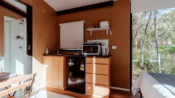 Eine kompakte Küche mit Mikrowelle, Wasserkocher und kleinem Kühlschrank. Es gibt eine hölzerne Sitzbank und ein Bett mit weißer Tagesdecke.
