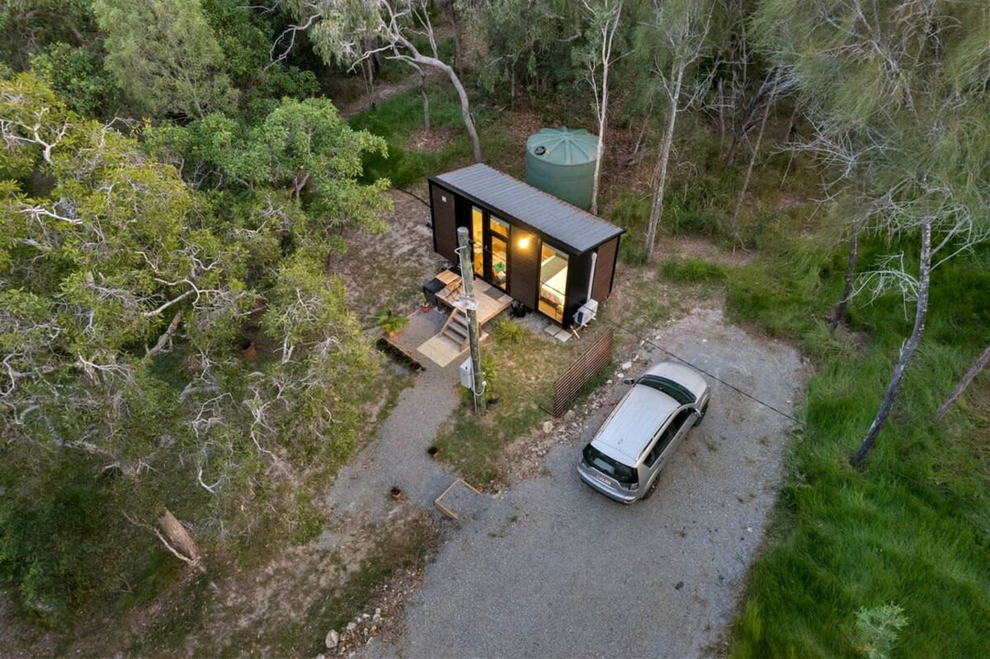 Une cabane moderne dotée d’une terrasse, avec une voiture garée devant, le tout entouré d’une forêt dense.