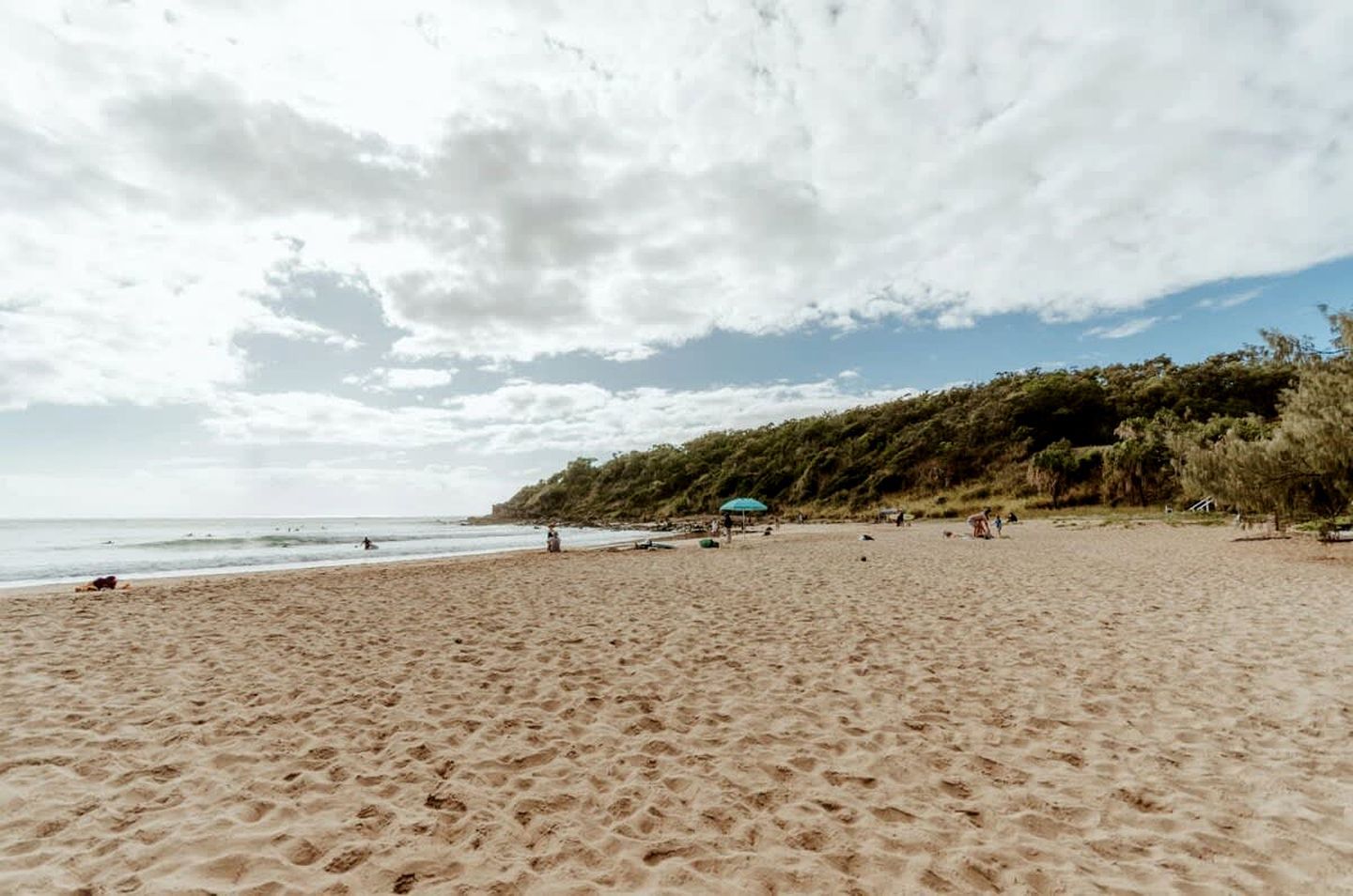 Praia de areia com céu claro e colina arborizada ao fundo.