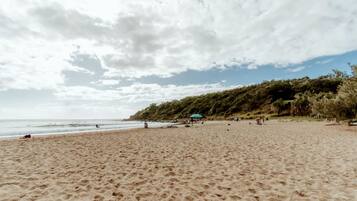 Praia de areia com céu claro e colina arborizada ao fundo.