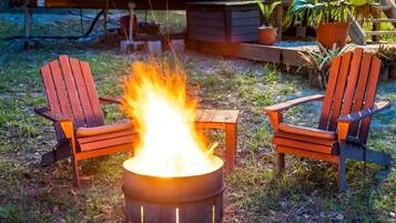 Een kampvuur met twee houten stoelen ervoor, gelegen op een grasveld met potplanten op de achtergrond.
