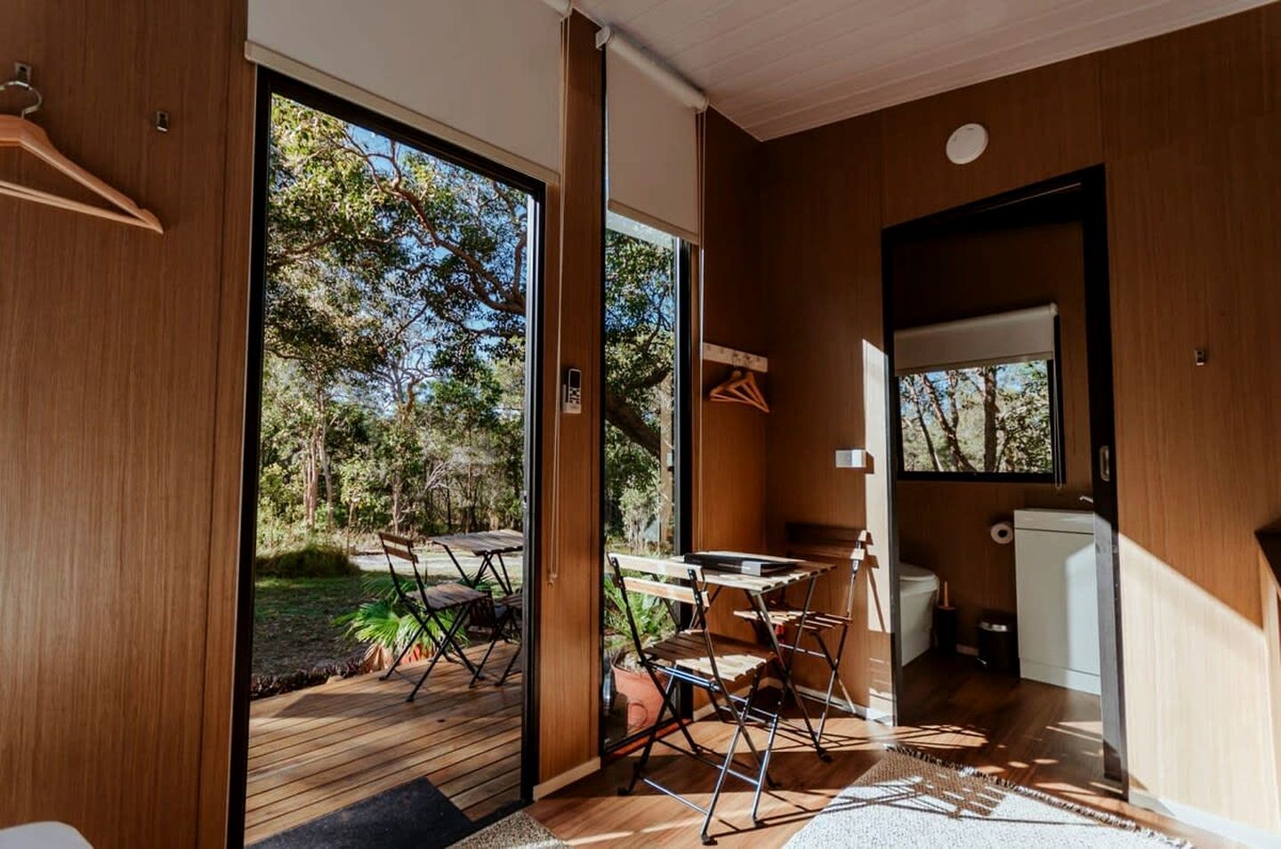 A modern interior with a wooden floor, a dining area with a table and chairs, a kitchen area with a refrigerator, and a large sliding glass door leading to an outdoor space with a deck and greenery.