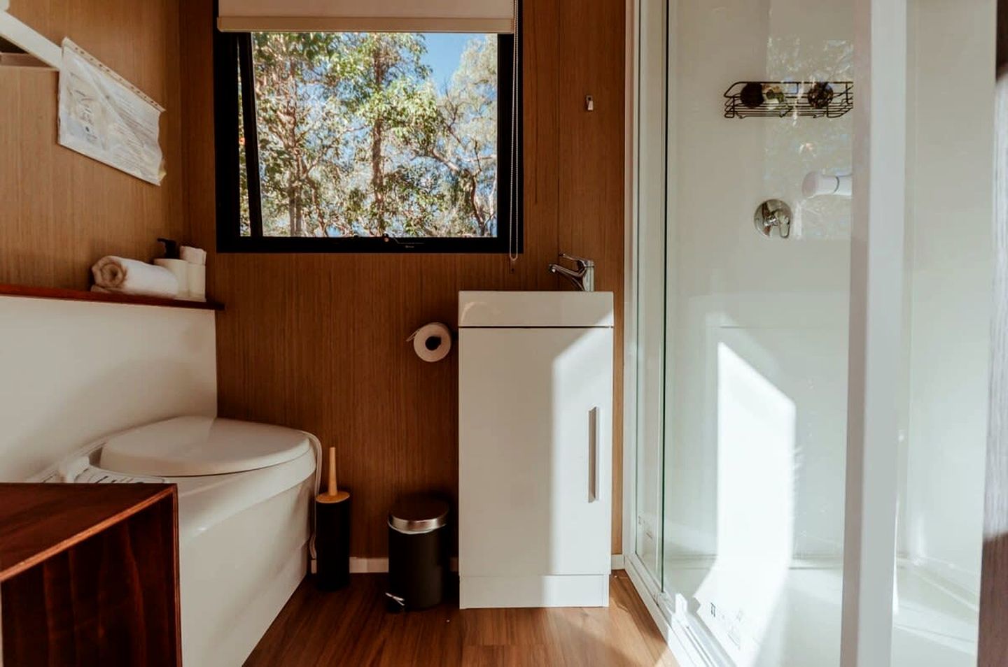 A compact bathroom with a toilet, a small sink, a window with a view of trees, and a glass shower enclosure.