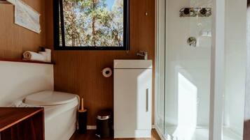 A compact bathroom with a toilet, a small sink, a window with a view of trees, and a glass shower enclosure.
