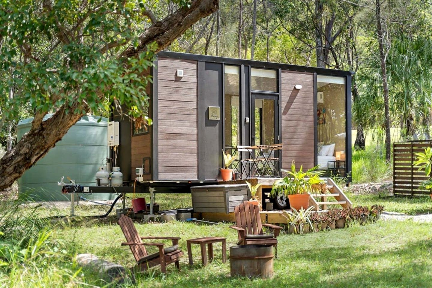 Una cabaña moderna y compacta con puerta de vidrio, rodeada por un jardín exuberante y mobiliario exterior.