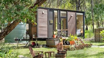Una cabaña moderna y compacta con puerta de vidrio, rodeada por un jardín exuberante y mobiliario exterior.