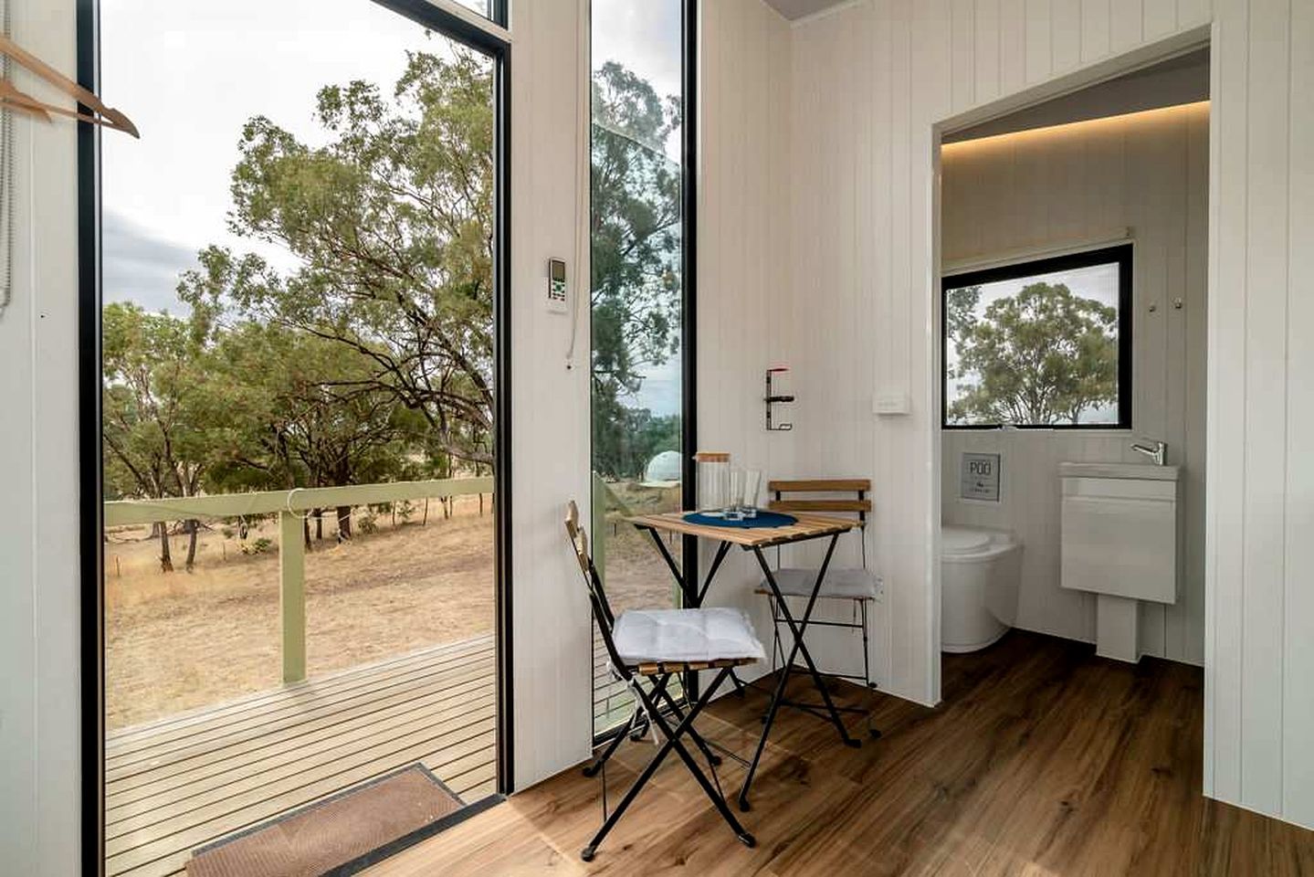 A room with a wooden floor, a small table with chairs, a window, and a bathroom with a sink and toilet.
