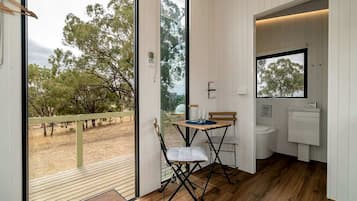 A room with a wooden floor, a small table with chairs, a window, and a bathroom with a sink and toilet.
