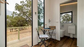 A room with a wooden floor, a small table with chairs, a window, and a bathroom with a sink and toilet.