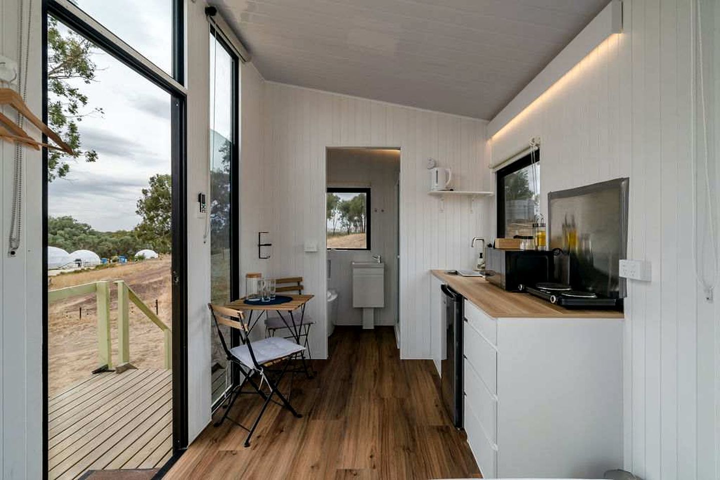 A compact kitchen with a wooden floor, a dining area with a table and chairs, and a large window offering a view of the outdoors.