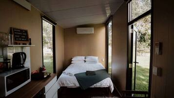 Una habitación individual con cama, escritorio con cafetera, un estante con objetos y vistas al exterior a través de amplias ventanas.