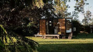A small, modern cabin with a wooden exterior, a door, and a window, situated in a grassy area with trees in the background.