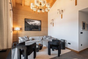 Ein modernes Wohnzimmer mit einem Ecksofa, einem Couchtisch und einem Fernseher. Es gibt eine Holzbalkendecke mit einem Kronleuchter, einer Wandlampe und einem gerahmten Bild.
