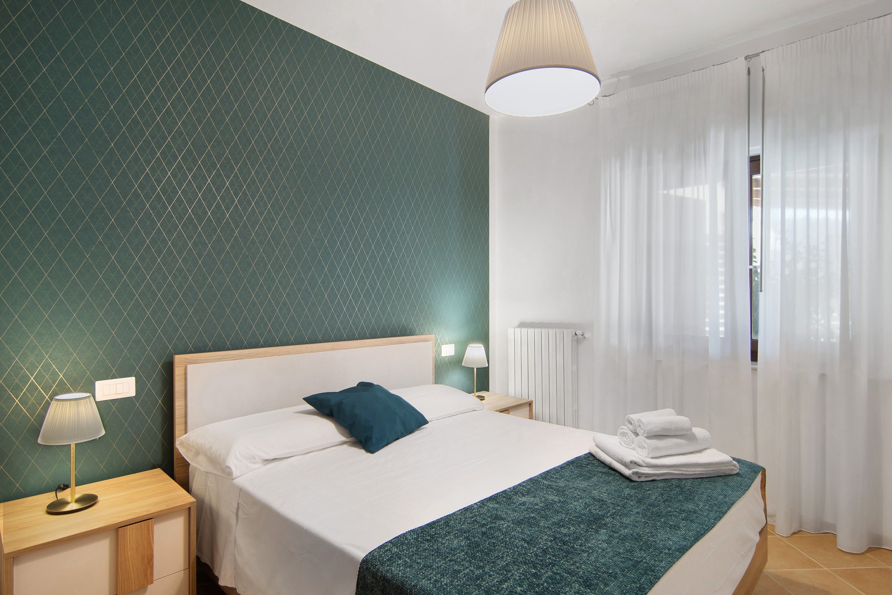 Habitación de hotel con cama, mesita de noche, lámpara, radiador y ventana con cortinas.