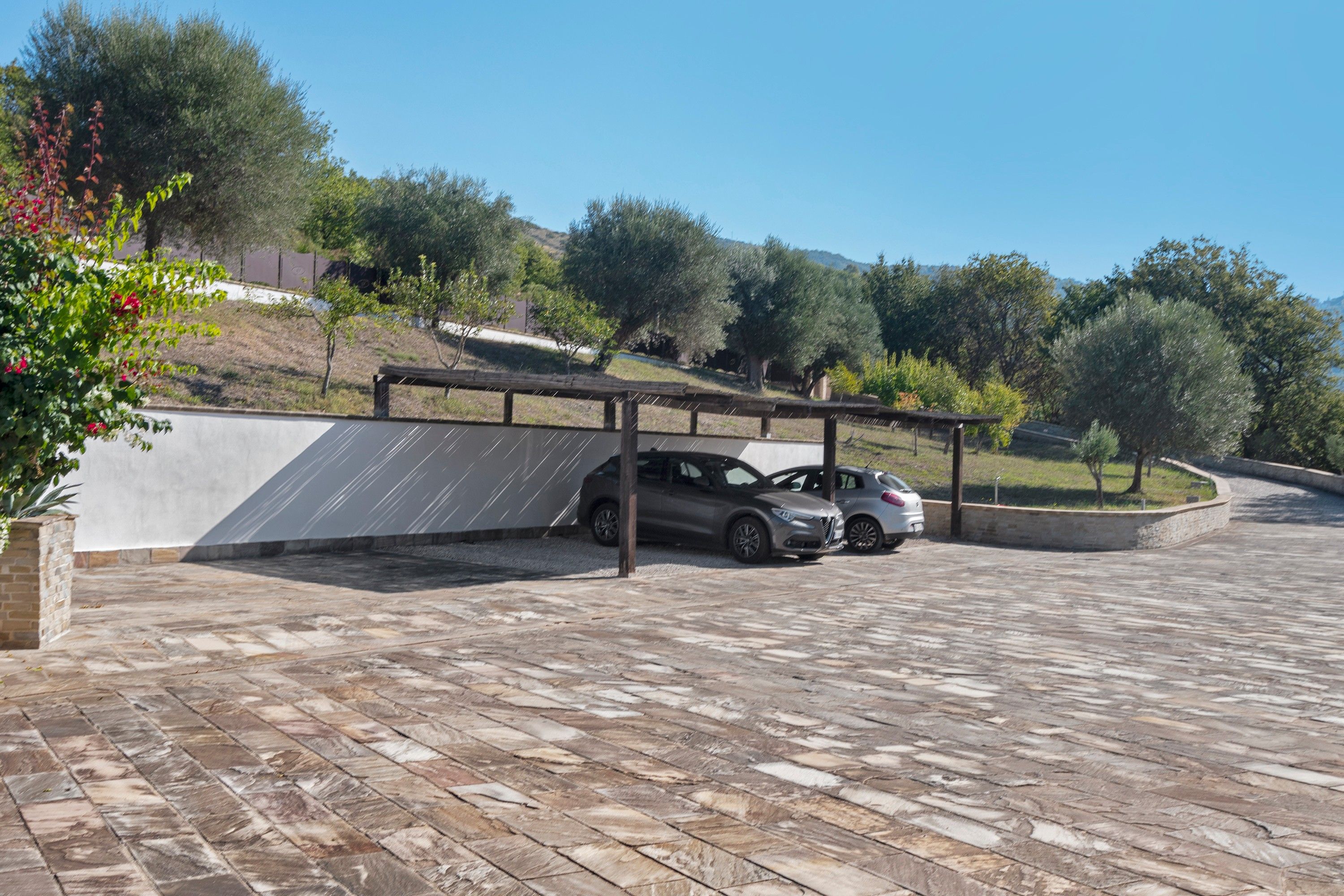 Un área de estacionamiento con dos coches aparcados bajo una estructura cubierta, rodeada de árboles y un paisaje montañoso.