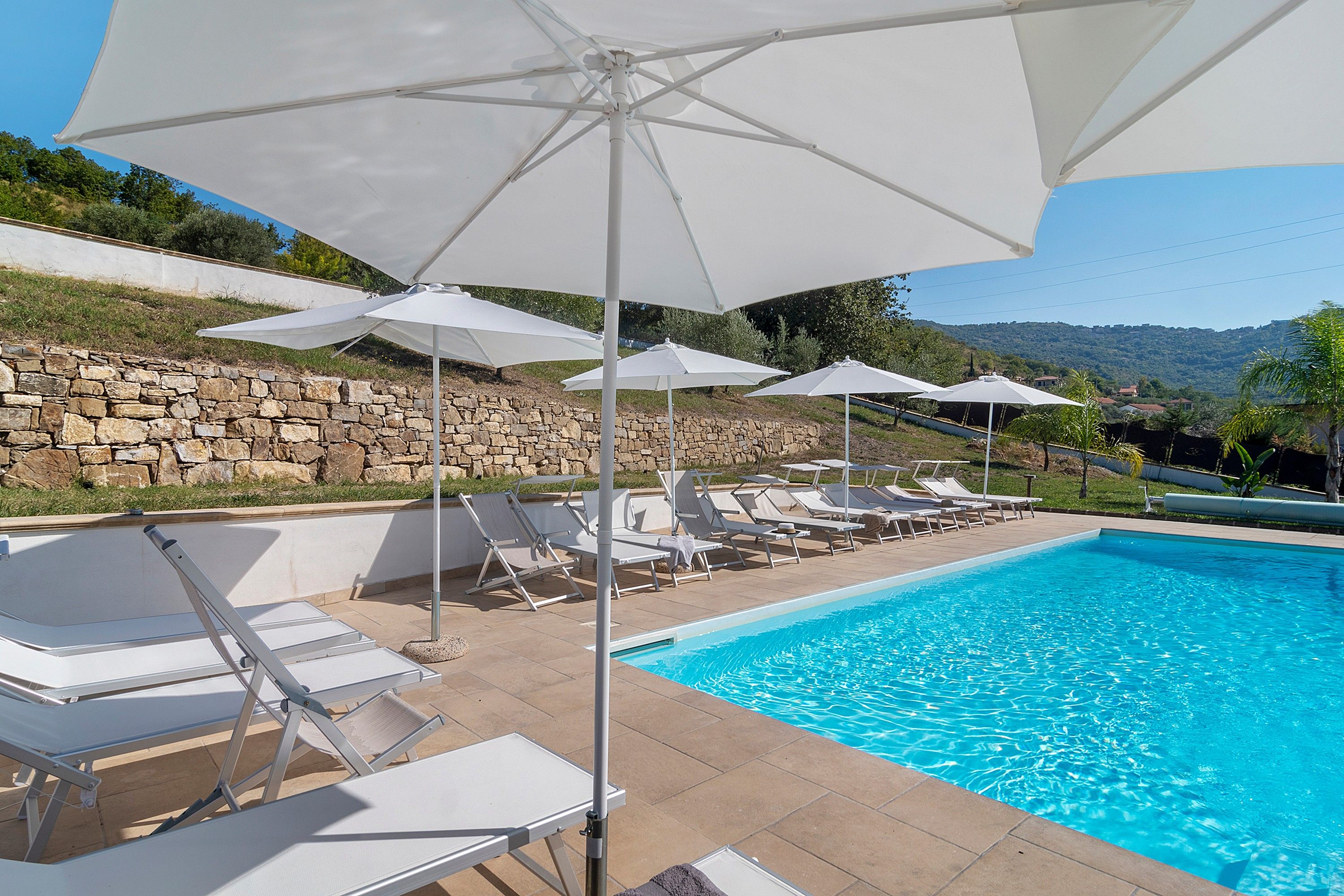 Una piscina con sillones blancos y sombrillas, rodeada de muros de piedra y follaje.