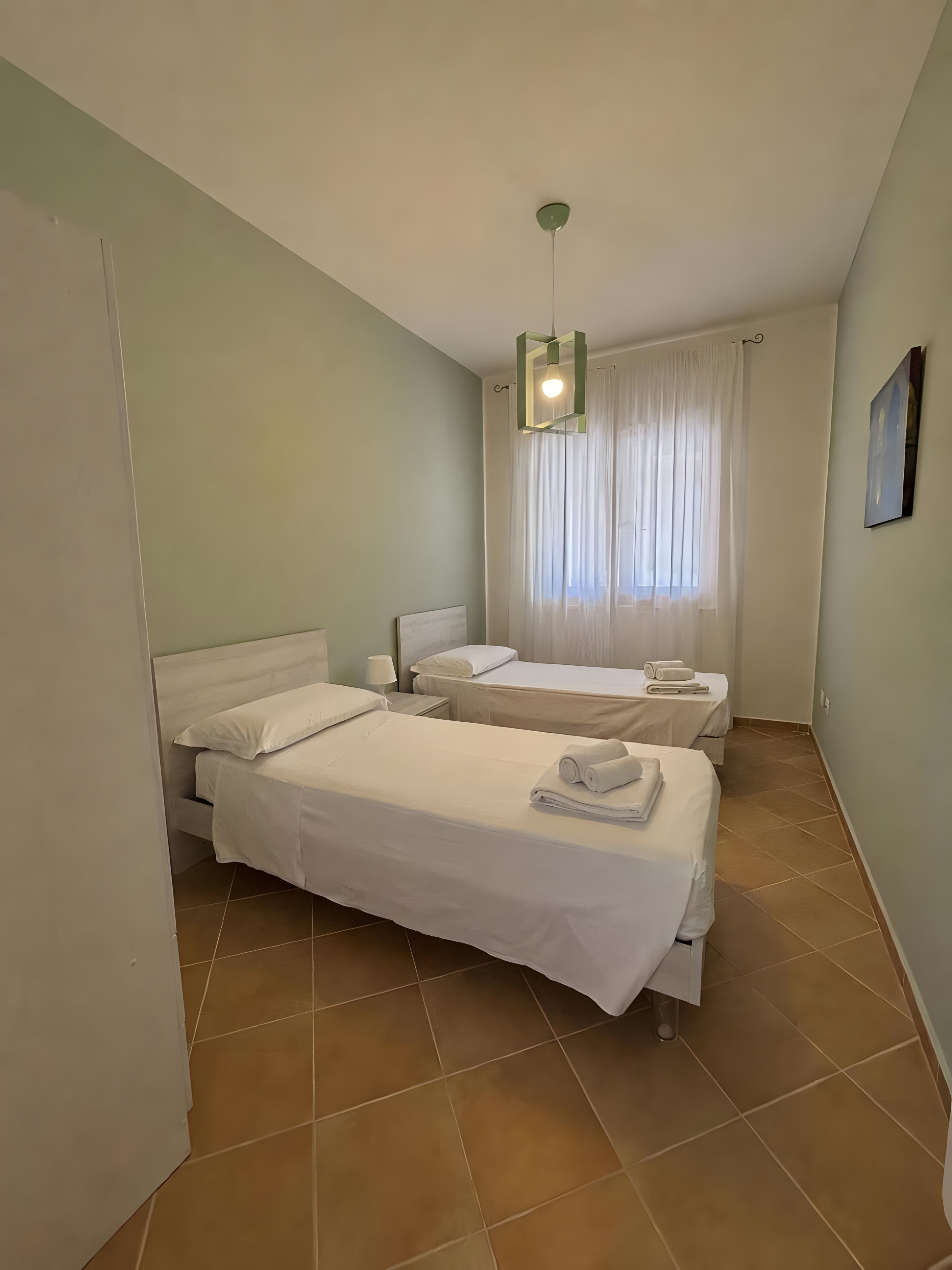 Habitación de hotel con dos camas, una lámpara en el techo, un ventanal con cortinas y suelo de baldosas.