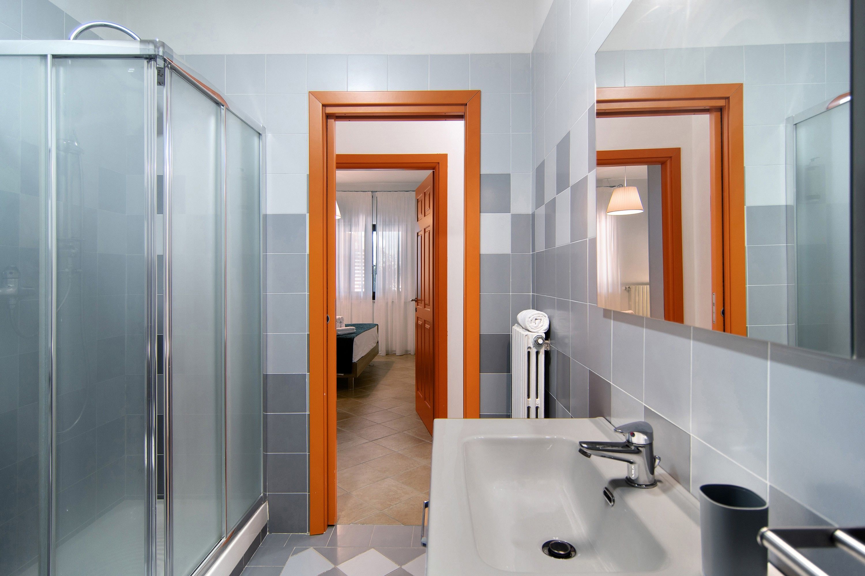 A modern bathroom with a glass shower enclosure, a sink with a mirror, and a hallway leading to a bedroom.