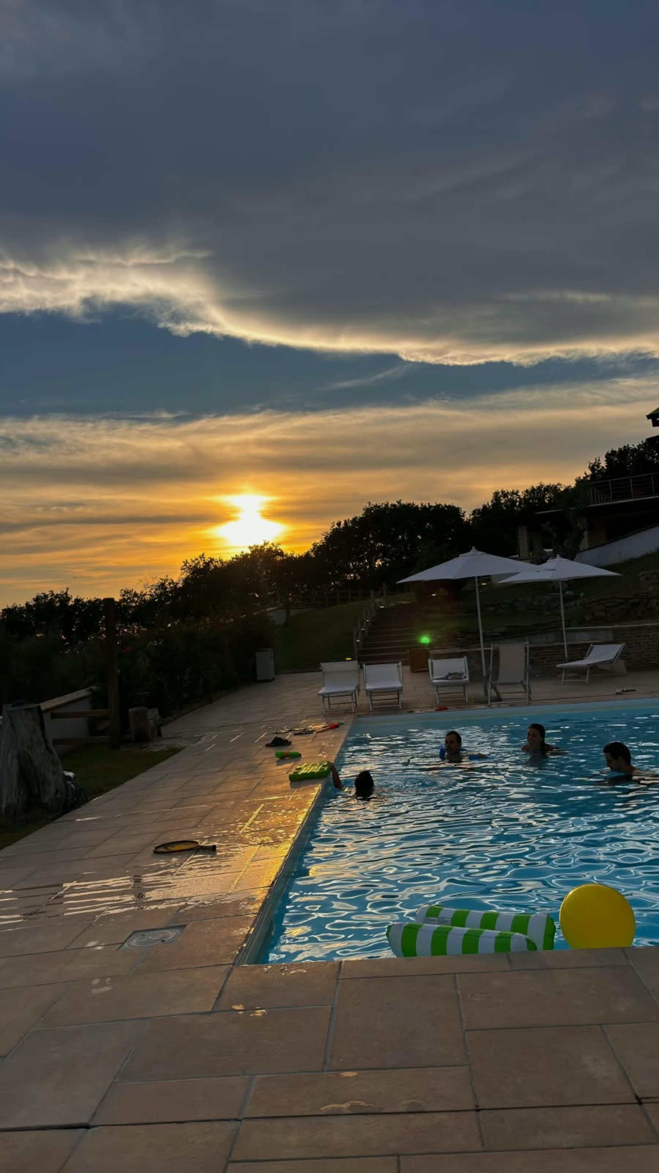 Una piscina con persone a nuotare, circondata da lettini e ombrelloni, con un tramonto sullo sfondo.