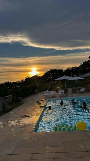 Una piscina con persone a nuotare, circondata da lettini e ombrelloni, con un tramonto sullo sfondo.