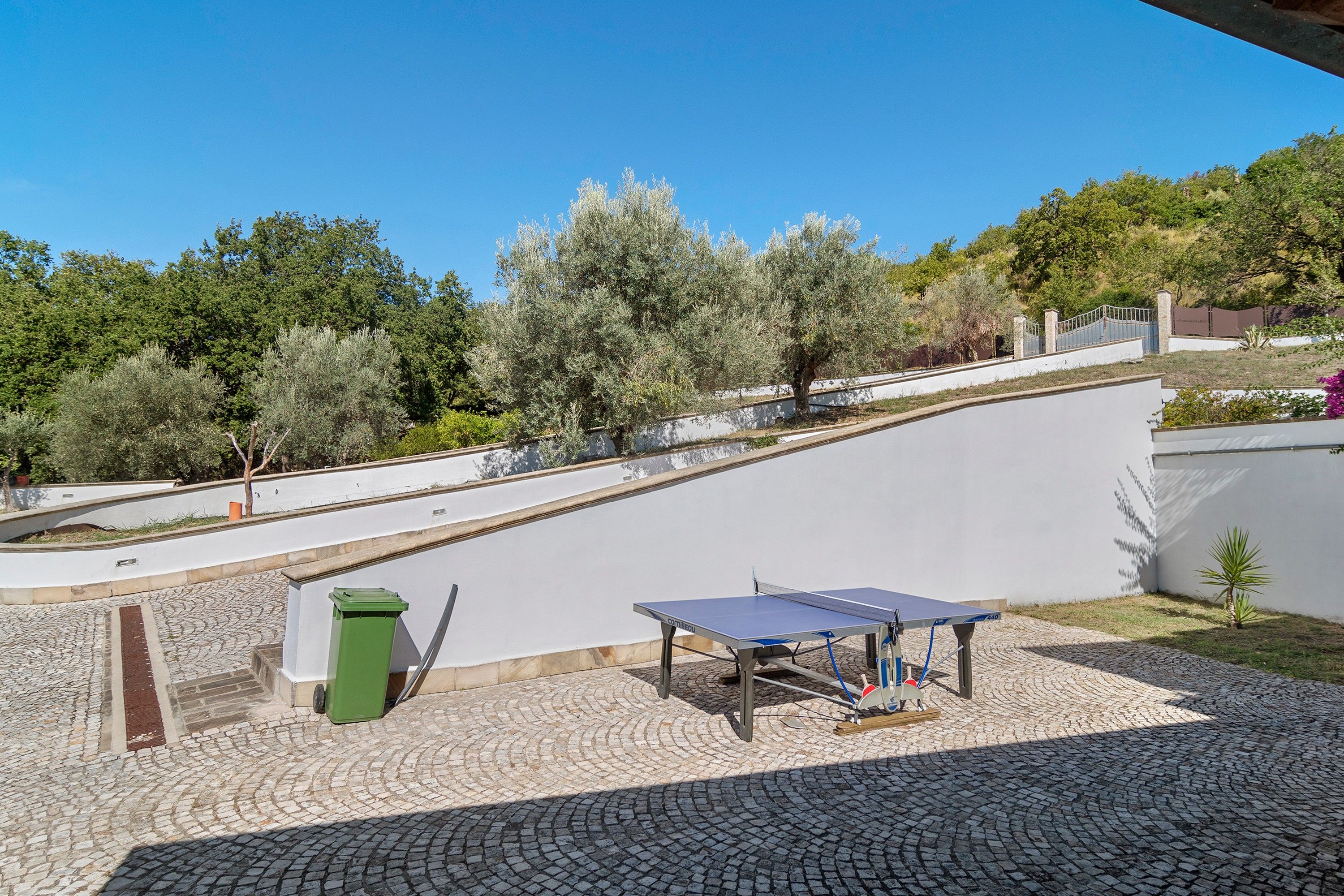 Un patio con mesa de ping pong, un bote de basura verde y una zona pavimentada.