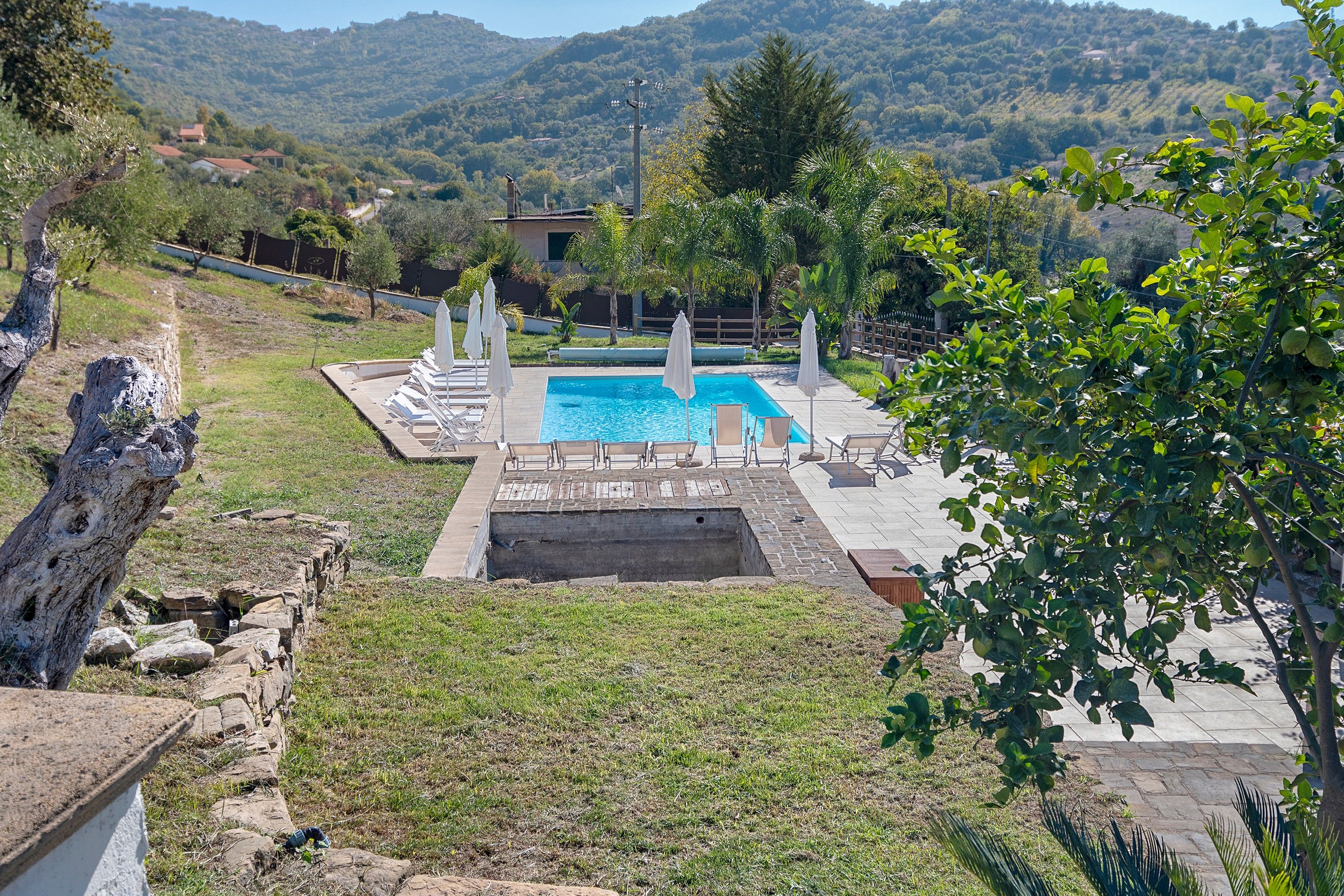 A swimming pool with lounge chairs and umbrellas, surrounded by a stone wall and greenery, with a mountainous backdrop.