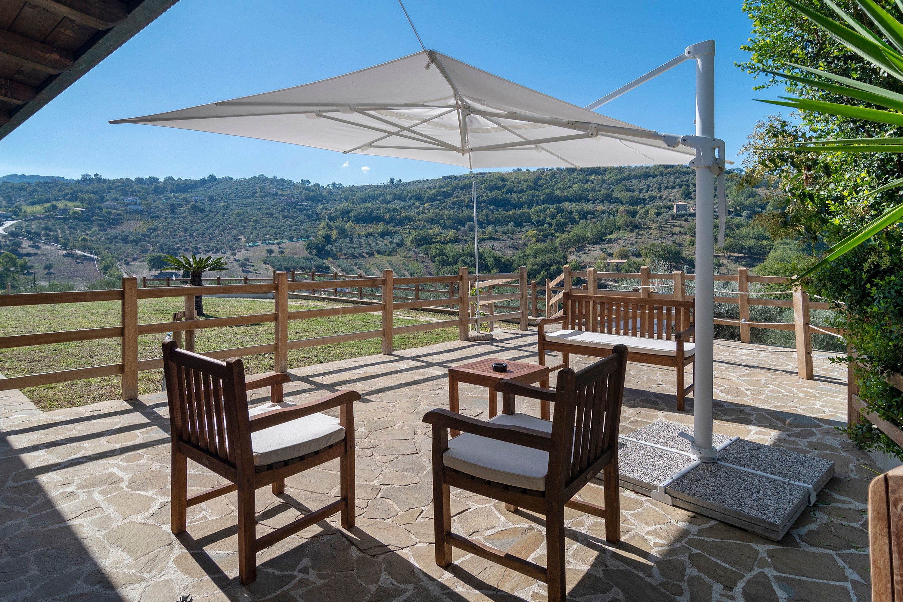 Eine Terrasse im Freien mit Steinböden, Holzmöbeln und einem großen Sonnenschirm, der Schatten spendet.