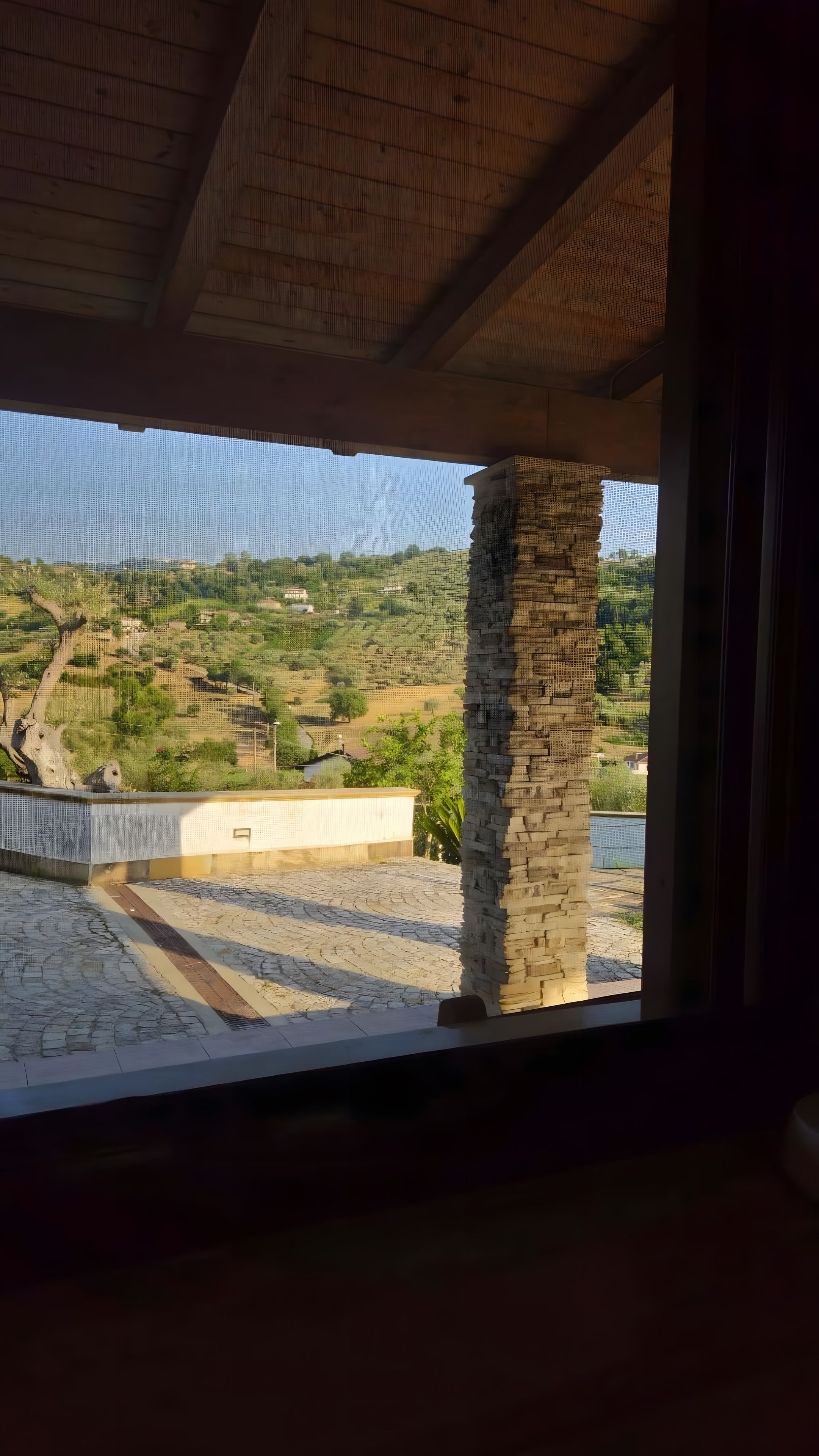 Una stanza con vista su un paesaggio collinare, visibile attraverso una finestra dotata di zanzariera.