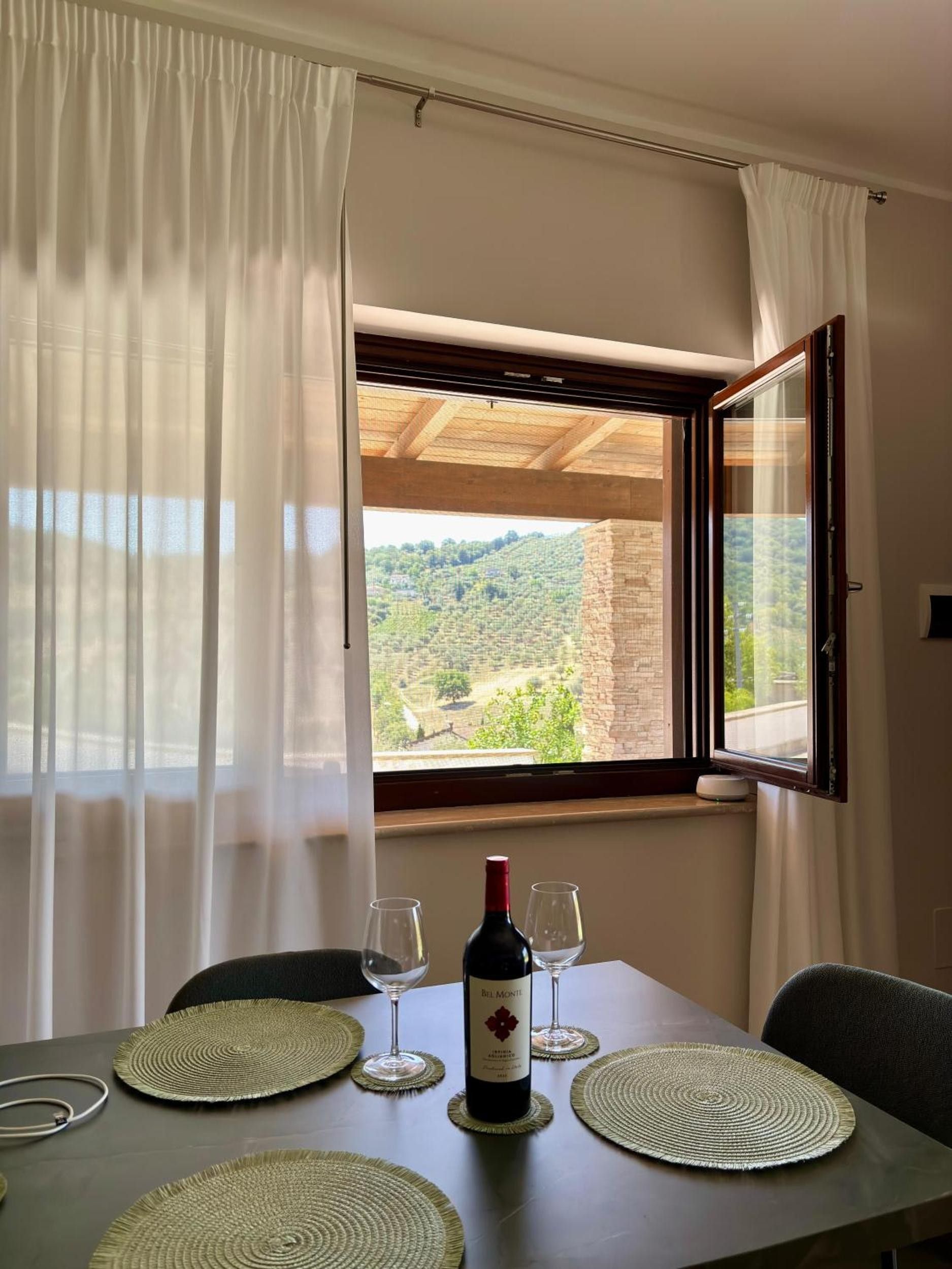 Un tavolo da pranzo con una bottiglia di vino e bicchieri, vista su un vigneto attraverso una finestra, e un soffitto in legno.