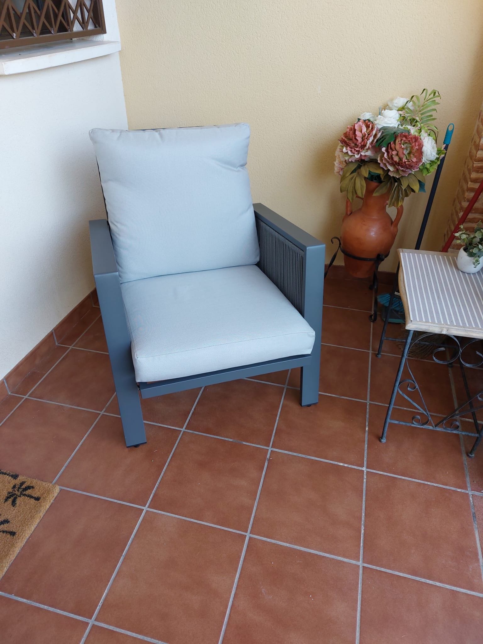 Una silla azul claro con cojín, una mesita con mantel a rayas y una maceta de terracota grande con flores.