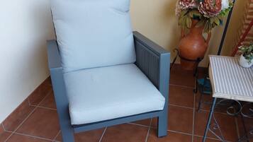 Una silla azul claro con cojín, una mesita con mantel a rayas y una maceta de terracota grande con flores.