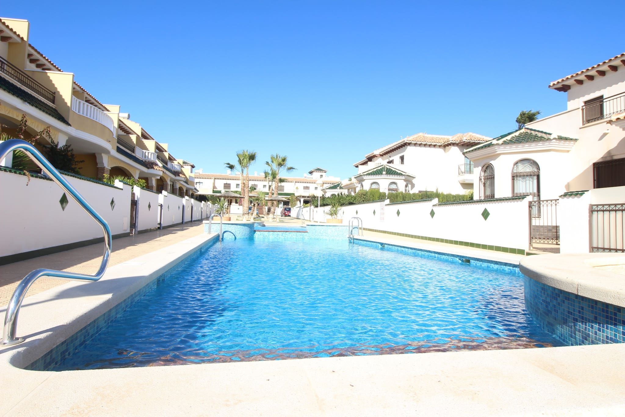 A swimming pool surrounded by multi-story buildings with balconies, a clear blue sky, and palm trees.