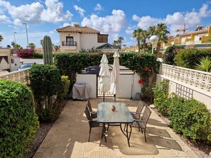 A patio with a table and chairs, surrounded by greenery and a building with a tiled roof. - Townhouse Maribel Cuidad Quesada (Rojales)
