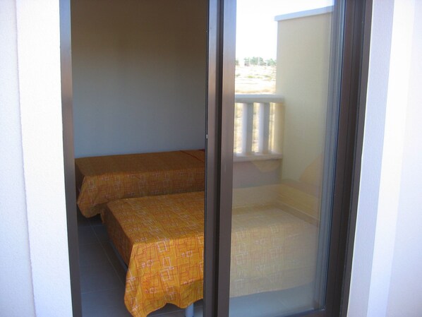 A room with two beds covered in patterned bedding, a window with a view of a building, and a balcony with a railing.