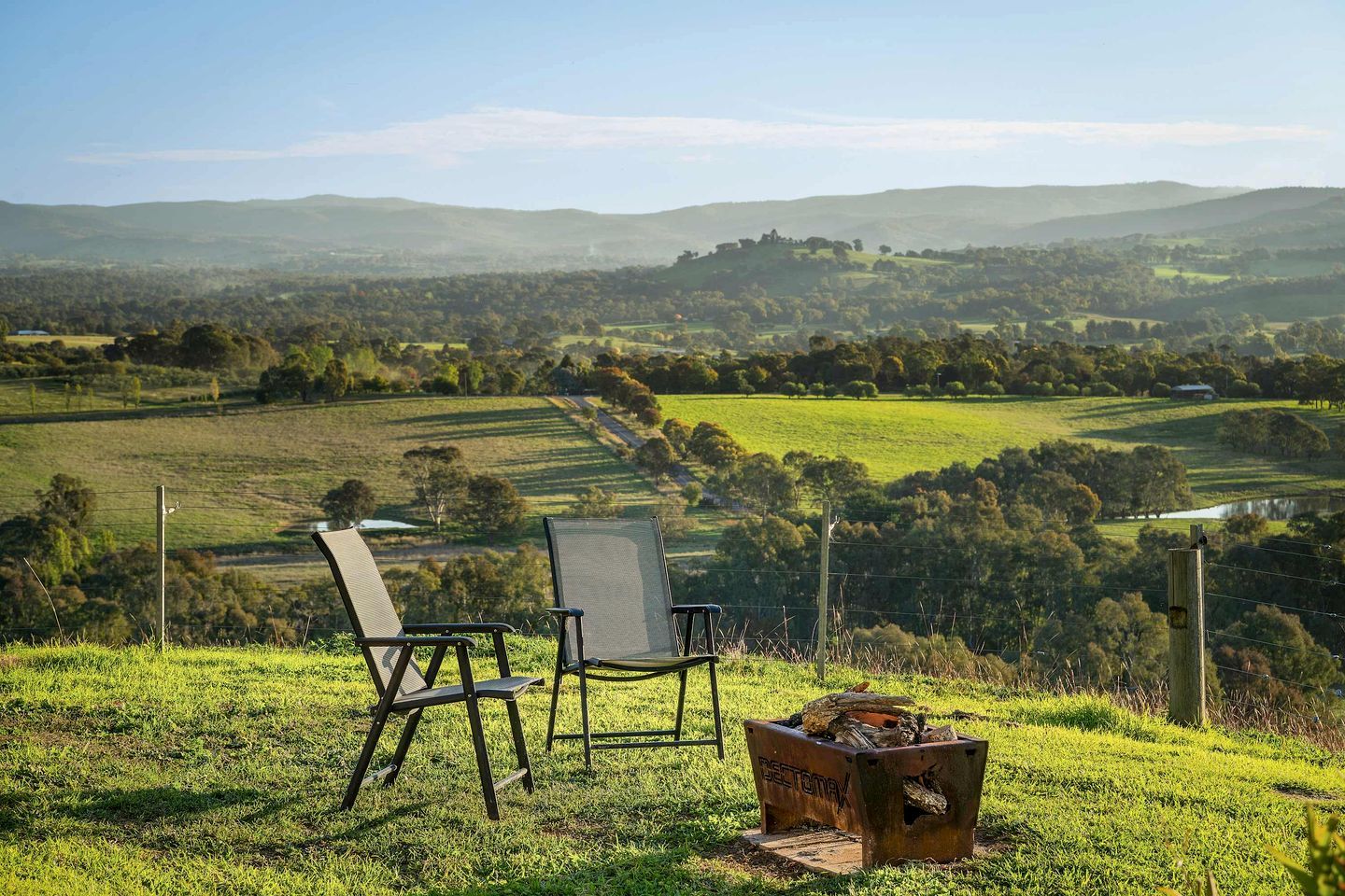 Un espace extérieur verdoyant, avec deux chaises et un poêle à bois, donnant sur une vallée parsemée de champs et bordée de collines lointaines.