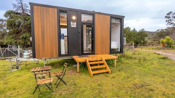 Una cabaña moderna y compacta con fachada de madera y ventanas amplias, ubicada en un área verde con terraza de madera y mobiliario.