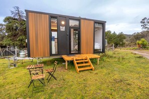 A modern, compact cabin with a wooden exterior and large windows, situated on a grassy area with a wooden deck and seating.