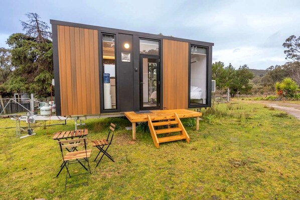 A modern, compact cabin with a wooden exterior and large windows, situated on a grassy area with a wooden deck and seating.