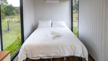Un dormitorio con una cama grande, ropa de cama blanca y vistas a un área verde por la ventana.