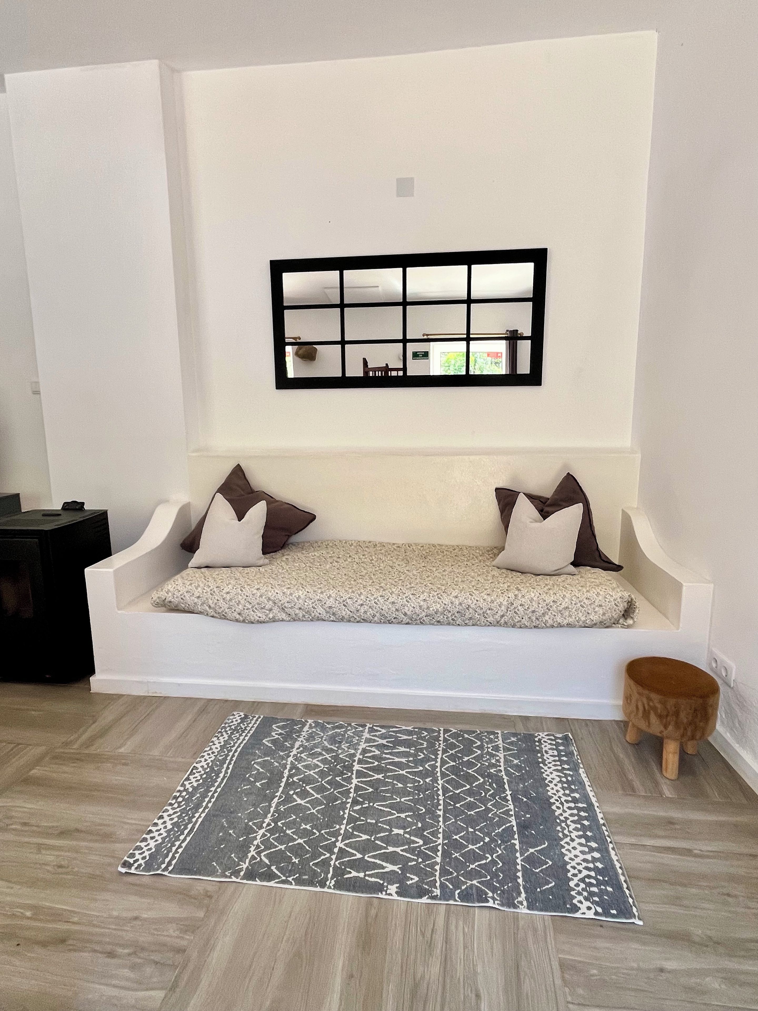 Een witte bank met twee kussens, een kamerkleed met patroon, en een klein houten krukje in een ruimte met een witte muur en een spiegel met een zwarte lijst.