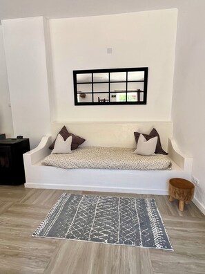 A white sofa with two pillows, a patterned rug, and a small wooden stool in a room with a white wall and a black-framed mirror.