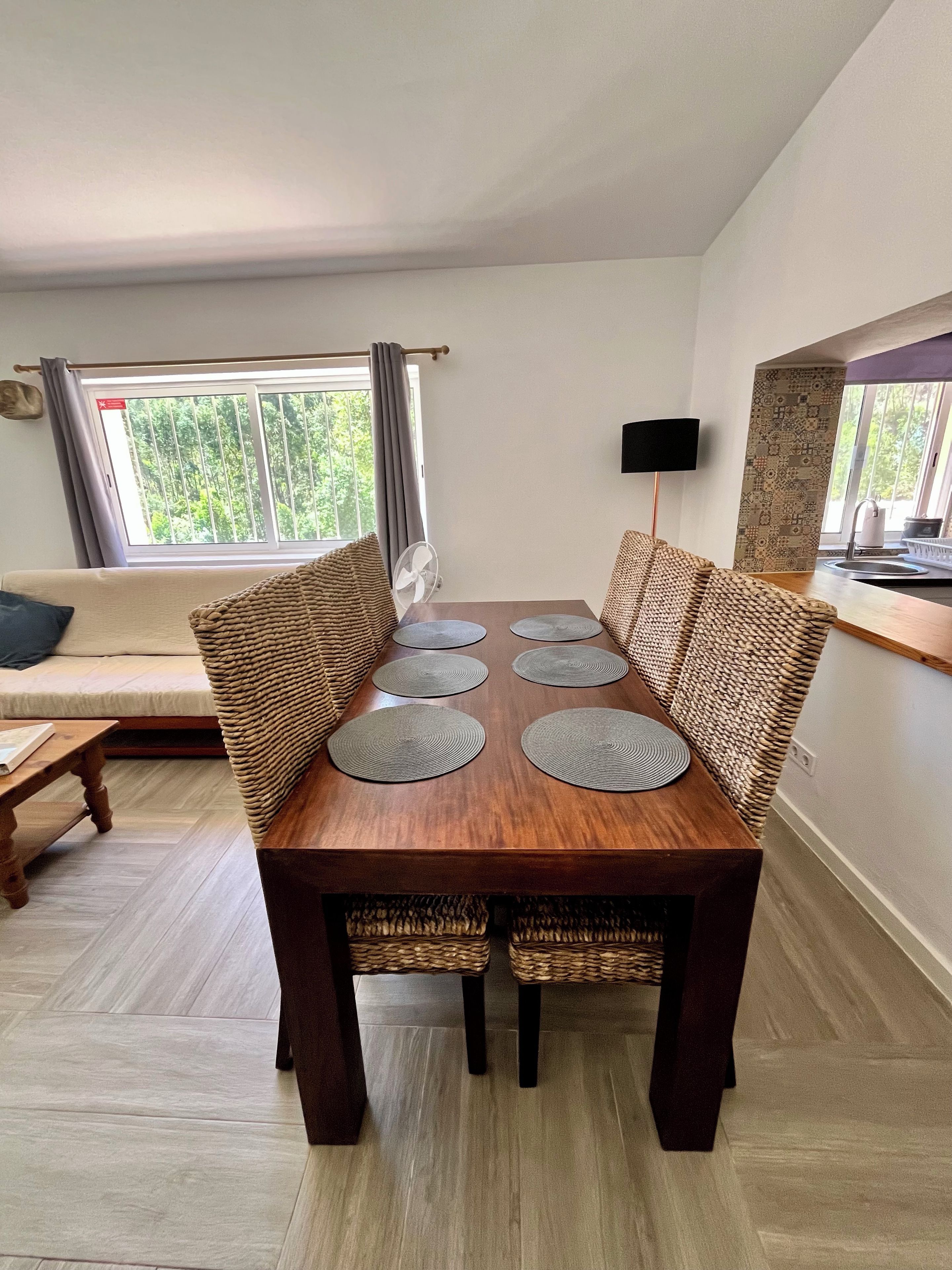 Un coin repas avec une table en bois, des chaises en osier et une vue sur la verdure par la fenêtre.