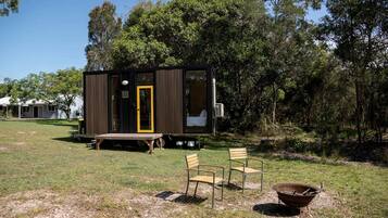 A small cabin with a door, a bench, two chairs, and a fire pit in a grassy area with trees.