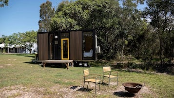 Una pequeña cabaña con puerta, un banco, dos sillas y un brasero en un área cubierta de césped y árboles.