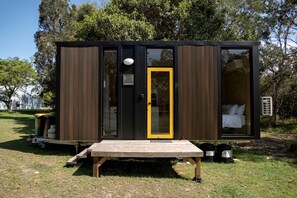 A small, modern cabin with a wooden deck, a single door with a yellow frame, and a visible bed inside.